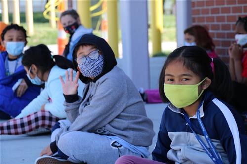 Two students look at the camera wearing face coverings and waving